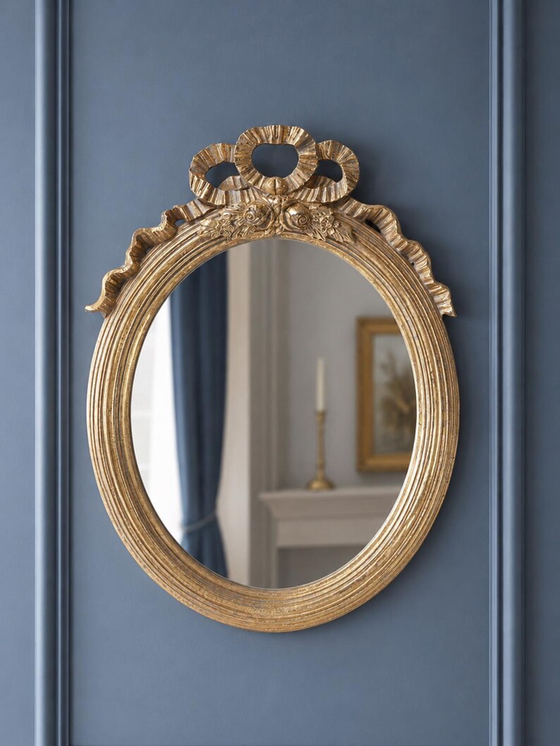 May include: Oval gold-colored mirror with a decorative ribbon and floral design at the top. The mirror has a ridged frame and is mounted on a blue wall. The reflection shows a glimpse of a room with a candle.