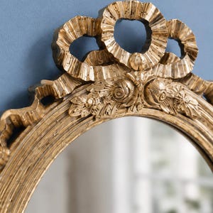 May include: Ornate gold-colored oval mirror with a decorative bow at the top. The frame features intricate floral and scrollwork details. The mirror's surface reflects a blurred interior scene, adding depth to the design. A classic piece for home decor.