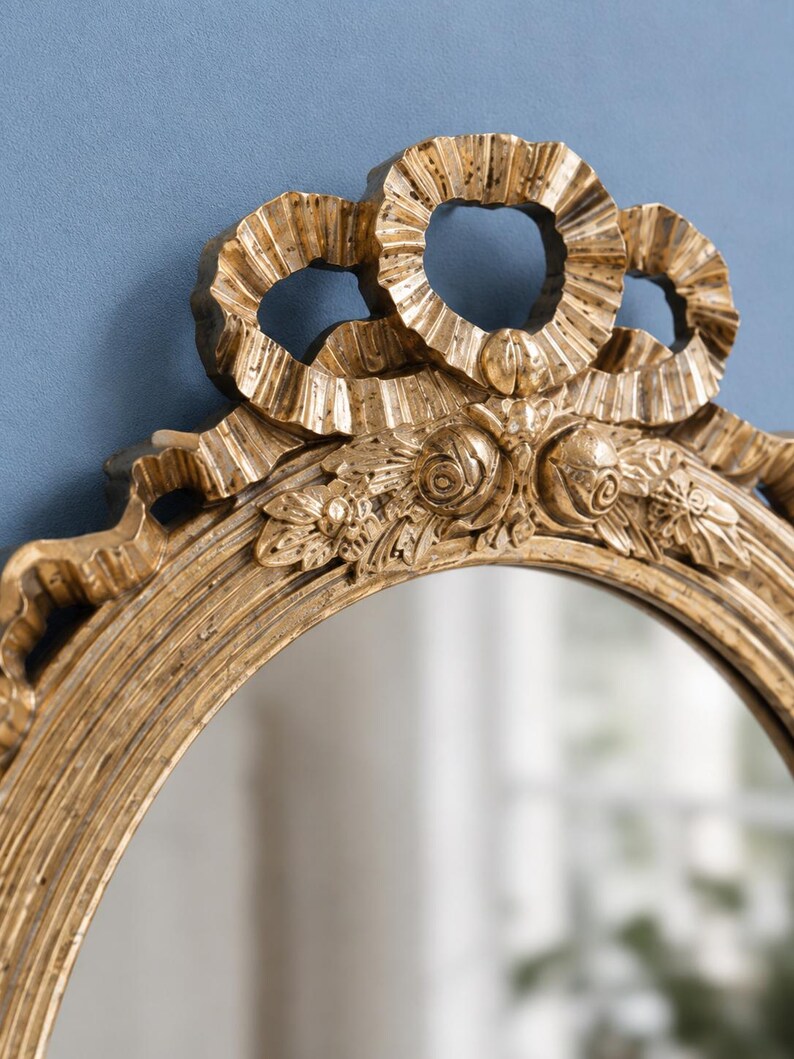 May include: Ornate gold-colored oval mirror with a decorative bow at the top. The frame features intricate floral and scrollwork details. The mirror's surface reflects a blurred interior scene, adding depth to the design. A classic piece for home decor.