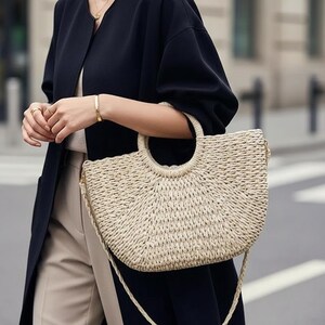 May include: A beige woven handbag with a rounded top handle and a long shoulder strap. The bag has a semi-circular shape and a textured surface. The bag is being held by a person wearing a black coat and gold jewelry.