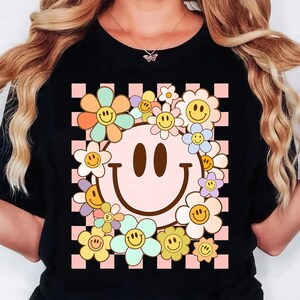 May include: Black t-shirt featuring a retro smiley face design. The smiley face is surrounded by colorful flowers with smiling faces. The background is a pink and white checkered pattern.