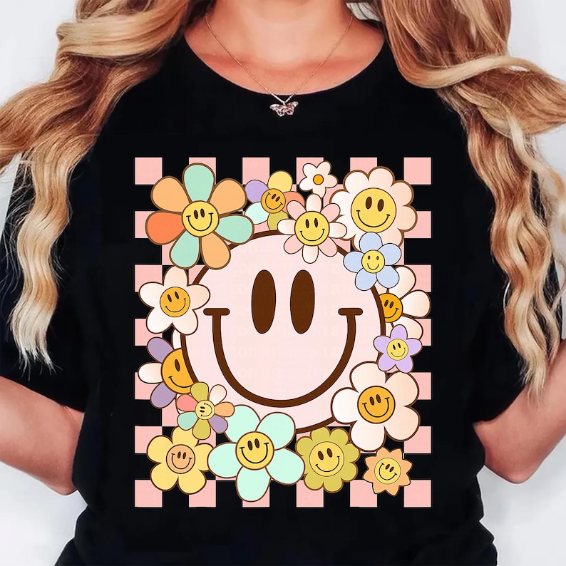 May include: Black t-shirt featuring a retro smiley face design. The smiley face is surrounded by colorful flowers with smiling faces. The background is a pink and white checkered pattern.