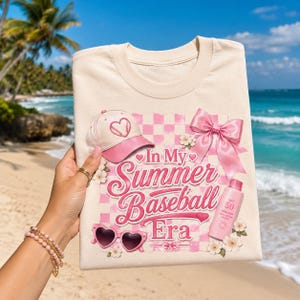 May include: Cream-colored t-shirt with a pink and white checkered design. The shirt features a baseball cap, heart-shaped sunglasses, a bow, and the text "In My Summer Baseball Era". A bottle of SPF 50 sunscreen is also visible.