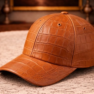 Op de afbeelding: Bruine baseballcap met een getextureerd, krokodilachtig patroon. De cap heeft een gebogen klep en een knoop aan de bovenkant. De hoed is gemaakt van een leerachtig materiaal en ligt op een lichtgekleurd tapijt.