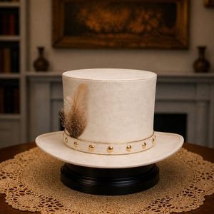 May include: A cream-colored top hat with a flat crown and a wide brim. The hat features a band around the base adorned with small, evenly spaced gold-toned metal studs and a single brown and cream-colored speckled feather tucked into the side.