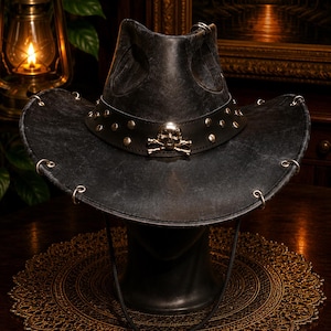 May include: A black distressed cowboy hat featuring a wide brim with silver-toned metal rings along the edge. A black leather band encircles the crown, adorned with silver-toned studs and a prominent metallic skull and crossbones emblem at the front.