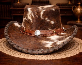 May include: A brown and white cowhide cowboy hat with a leather band and decorative silver-colored accents. The hat has a stitched brim and is displayed on a round, woven placemat.