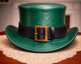 Peut inclure: Un chapeau haut de forme en cuir vert avec une bande noire et une boucle dorée. Le chapeau a un large bord et est posé sur une table en bois avec un napperon en dentelle décoratif.