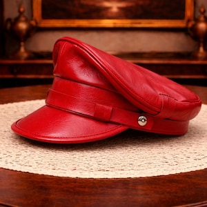 Pode incluir: Um chapéu de couro vermelho brilhante com viseira, faixa e detalhe de botão. O chapéu está sobre uma toalha de mesa circular creme em uma mesa de madeira escura. O fundo inclui uma pintura e uma estante.