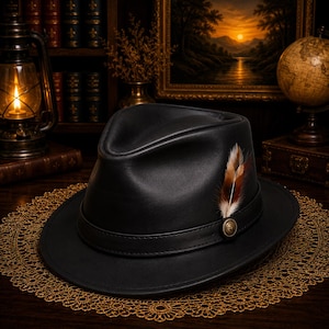 Könnte beinhalten: Ein schwarzer Fedora-Hut mit glatter Oberfläche, strukturierter Krone und breiter Krempe. Der Hut ist mit einem schwarzen Lederband, einem kleinen runden Metallknopf und einer braun-weiß gemusterten Feder an der Seite verziert.