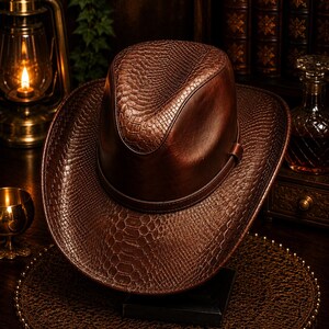 May include: A brown fedora hat featuring a textured snakeskin pattern on the brim and crown, accented by a smooth leather band encircling the base. The hat displays a deep, rich brown colour with a glossy finish and a structured, pinched crown design.