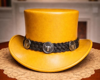 Peut inclure: Un haut-de-forme jaune moutarde doté d'une bande de chapeau en cuir tressé noir. La bande est ornée de trois conchos métalliques circulaires argentés, chacun gravé d'un motif détaillé de crâne de taureau centré sur le devant.