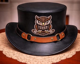 Peut inclure: Haut-de-forme en cuir noir avec un large bord et une bande en cuir marron. Le chapeau présente un écusson de hibou brodé avec des couleurs marron, beige et crème. Le chapeau est posé sur une table en bois.