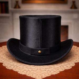 Könnte beinhalten: Ein schwarzer Zylinderhut mit breiter Krempe und schwarzem Band. Der Hut hat einen goldfarbenen Knopf am Band. Er liegt auf einem braunen Tisch mit einer dekorativen Spitzendecke.