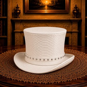 Puede incluir: Un sombrero de copa blanco con un patr&oacute;n texturizado y una banda con tachuelas descansa sobre un tapete marr&oacute;n adornado. El sombrero se exhibe sobre una mesa de madera oscura, con una chimenea y estanter&iacute;as en el fondo.