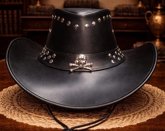 May include: Black leather cowboy hat with silver-tone studs and a skull and crossbones emblem on the band. The hat has a wide brim and a chin strap. The hat is sitting on a brown table.