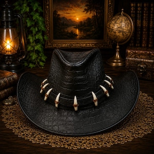 Peut inclure: Un chapeau de cowboy noir avec un motif texturé en peau de crocodile, doté d'une bande décorative ornée de plusieurs accents blancs incurvés en forme de dents, fixés par de petits anneaux couleur cuir brun autour de la calotte.