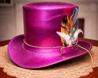Peut inclure: Un haut de forme magenta vif avec un aspect métallique. Le chapeau est orné d'une bande décorative et de plumes colorées. Un cordon doré entoure le chapeau. Le chapeau est présenté sur une table en bois.