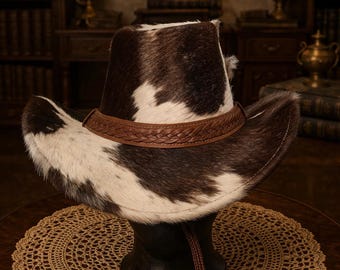 Puede incluir: Sombrero de vaquero con una banda de cuero marrón y un patrón de piel de vaca en tonos marrones y blancos. El sombrero tiene un ala ancha y una correa para la barbilla. El sombrero se exhibe sobre un soporte de madera oscura.
