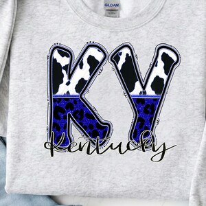 Peut inclure: Un sweat-shirt gris clair avec les lettres "KY" dans un motif de vache noir et blanc sur un imprimé léopard bleu. Le mot "Kentucky" est écrit en cursive en dessous. Le sweat-shirt est plié à côté d'un jean bleu.