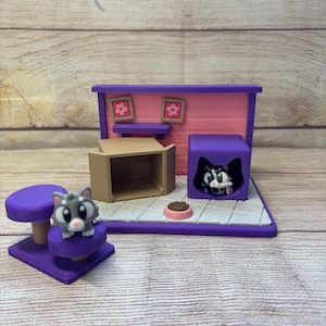 Puede incluir: Un juego de gatos en miniatura con una base morada, una casa rosa y morada y una caja de cartón. El juego incluye una figura de gato gris sobre una percha morada y una figura de gato blanco y negro en un cubo morado.