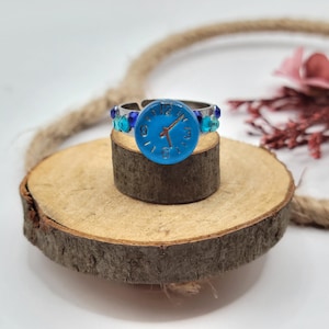May include: A silver-toned ring with a blue clock face design. The ring features a round, light blue clock face with black hands and numbers. The band is adorned with blue and teal beads. The ring is displayed on a small wooden slice.