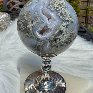 May include: A decorative agate sphere with a geode centre, displayed on a silver-coloured metal stand. The sphere shows blue, grey, and white patterns. The stand has a round base and a detailed stem. The sphere is approximately 15 cm in diameter.