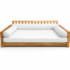 May include: A wooden daybed with a white mattress and two white bolster pillows. The frame is made of light brown wood. The back of the daybed has vertical wooden slats.