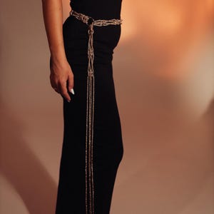 May include: A metallic gold-toned rope belt featuring a braided design with a circular ring buckle and long, dangling strands that extend downward. The belt is worn over a solid black garment, highlighting the contrast between the gold and black.