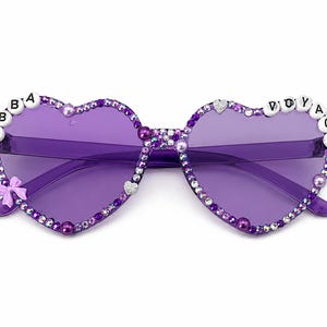 Könnte beinhalten: Herzförmige Sonnenbrille mit violett getönten Gläsern, deren Rahmen mit violetten und klaren Strasssteinen, kleinen weißen Buchstabenperlen mit der Aufschrift ABBA VOYAGE, einer kleinen hellvioletten Schleife und Herzakzenten verziert ist.