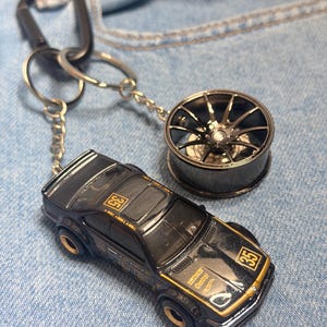 Puede incluir: Llavero de coche en miniatura negro y dorado con un dije de rueda a juego. El coche tiene el número 35 y el texto "Castrol". El llavero está sujeto a un mosquetón negro.