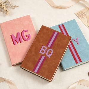 Peut inclure: Trois carnets rectangulaires aux couvertures lisses rose, marron et bleu clair. Le carnet rose porte les lettres MG en rose et rouge. Le carnet marron présente une bande verticale avec les lettres BQ. Le carnet bleu a une bande et la lettre Y.