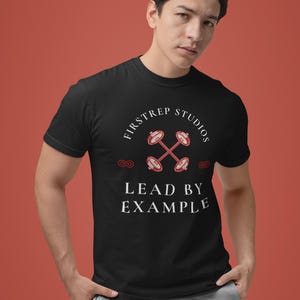 Peut inclure: T-shirt noir avec le texte "FIRSTREP STUDIOS" et "LEAD BY EXAMPLE" en blanc. Le t-shirt présente un motif d'haltères rouges et blancs. Le modèle porte un pantalon de survêtement gris.