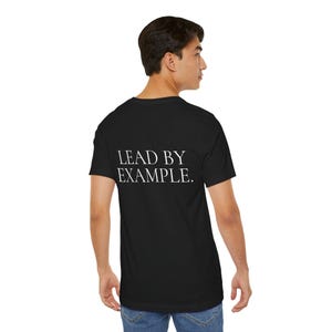 Peut inclure: T-shirt noir avec l'inscription blanche "LEAD BY EXAMPLE" au dos. Le t-shirt est de style col rond classique, en tissu doux. La personne porte un jean bleu.