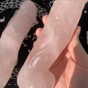 Könnte beinhalten: Zwei rosafarbene Rosenquarzskulpturen. Eine wird in einer Hand gehalten, die andere befindet sich im Hintergrund. Die Skulpturen sind glatt und poliert, mit einem leicht durchscheinenden Aussehen. Die Farbe ist ein weiches, natürliches Rosa.