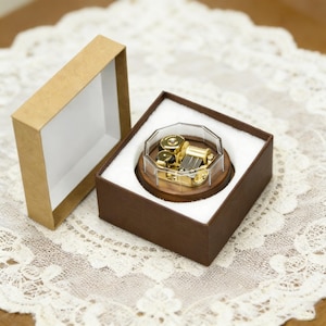 Puede incluir: Una pequeña caja de música dorada con una cubierta geométrica transparente se encuentra dentro de una caja marrón con interior blanco. La caja de música está sobre una base de madera. Una caja de cartón abierta y vacía está al lado.