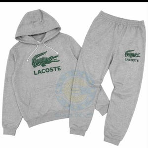Puede incluir: Un conjunto gris de sudadera con capucha y pantalón de chándal Lacoste. La sudadera con capucha presenta un logotipo de cocodrilo verde y el nombre de la marca Lacoste. El pantalón de chándal también tiene el logotipo de Lacoste. El conjunto está hecho de un material suave y cómodo.