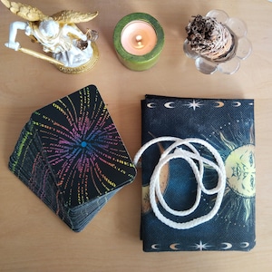 Può includere: Un mazzo di carte dei tarocchi con testo colorato e un punto blu al centro, accanto a un astuccio blu scuro con un disegno di sole e luna. Un cordoncino bianco è arrotolato sopra l'astuccio. Sono presenti anche una candela accesa in un supporto verde e una piccola statua di angelo.