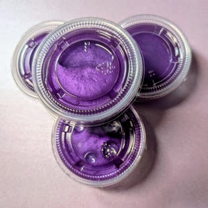 May include: Four small, clear plastic containers filled with a vibrant, shimmering purple substance. The containers have clear, snap-on lids. The purple material has a textured, almost granular appearance, catching the light and creating a sparkling effect.