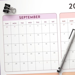 May include: A white paper calendar page for September with a black metal binder clip at the top left corner and a white fine liner pen with black text resting diagonally across the grid. The calendar features a grid layout with black lines.