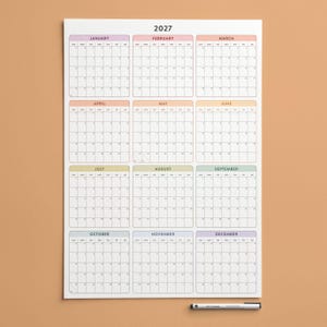 May include: A 2027 wall calendar printed on white paper, featuring twelve monthly grids arranged in a four-by-three layout. Each month is labelled with a distinct pastel-coloured header, and a black marker is positioned horizontally below the calendar.