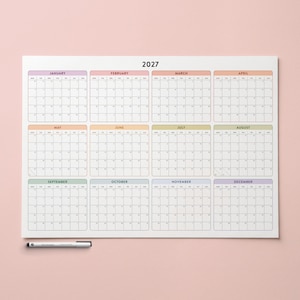 Könnte beinhalten: Ein Wandkalender für das Jahr 2027 mit zwölf Monatsübersichten in einem Vier-mal-drei-Layout. Jeder Monat ist von einem dünnen farbigen Rand umgeben und enthält die Wochentage. Ein schwarzer Stift liegt horizontal unter dem Kalender.