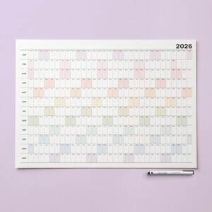 May include: A large, rectangular 2026 year planner with a white background and a grid layout. Each month is listed vertically, with days horizontally. The days are color-coded in pastel shades. A black and silver pen rests at the bottom.