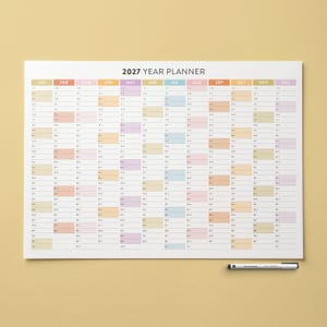 Puede incluir: Un planificador anual grande de 2027 con un fondo blanco y una cuadrícula mensual colorida. Los meses están etiquetados en la parte superior en varios colores. Un bolígrafo negro y plateado descansa en la esquina inferior derecha. El texto "2027 YEAR PLANNER" está en la parte superior.