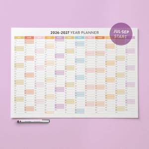 Può includere: Un grande pianificatore annuale 2026-2027 con un layout a griglia colorato. Il pianificatore include i mesi da luglio a marzo, con un'etichetta "Jul-Sep Start". Una penna nera è appoggiata in basso. Lo sfondo è viola chiaro.