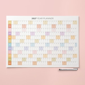 Pode incluir: Um grande planejador anual de 2027 com fundo branco e blocos pastel coloridos para cada dia. O texto "2027 YEAR PLANNER" está no topo. Uma caneta preta repousa no canto inferior direito.
