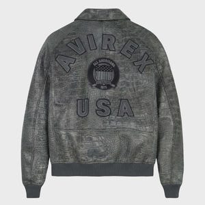 Puede incluir: Chaqueta de cuero gris con textura de cocodrilo. La chaqueta presenta la palabra "AVIREX" en los hombros y "USA" en la parte inferior. Un parche circular en el centro dice "ALL AMERICANS ESTABLISHED 1975".