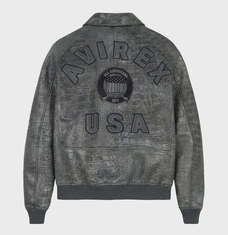 Puede incluir: Chaqueta de cuero gris con textura de cocodrilo. La chaqueta presenta la palabra "AVIREX" en los hombros y "USA" en la parte inferior. Un parche circular en el centro dice "ALL AMERICANS ESTABLISHED 1975".