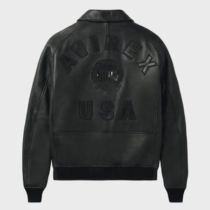 Puede incluir: Chaqueta de cuero negra con la palabra "AVIREX" arqueada en la parte superior de la espalda y "USA" debajo. La chaqueta tiene cuello, pu&ntilde;os y dobladillo de canal&eacute;. Un emblema circular est&aacute; centrado entre las palabras. La chaqueta es de estilo bomber cl&aacute;sico.