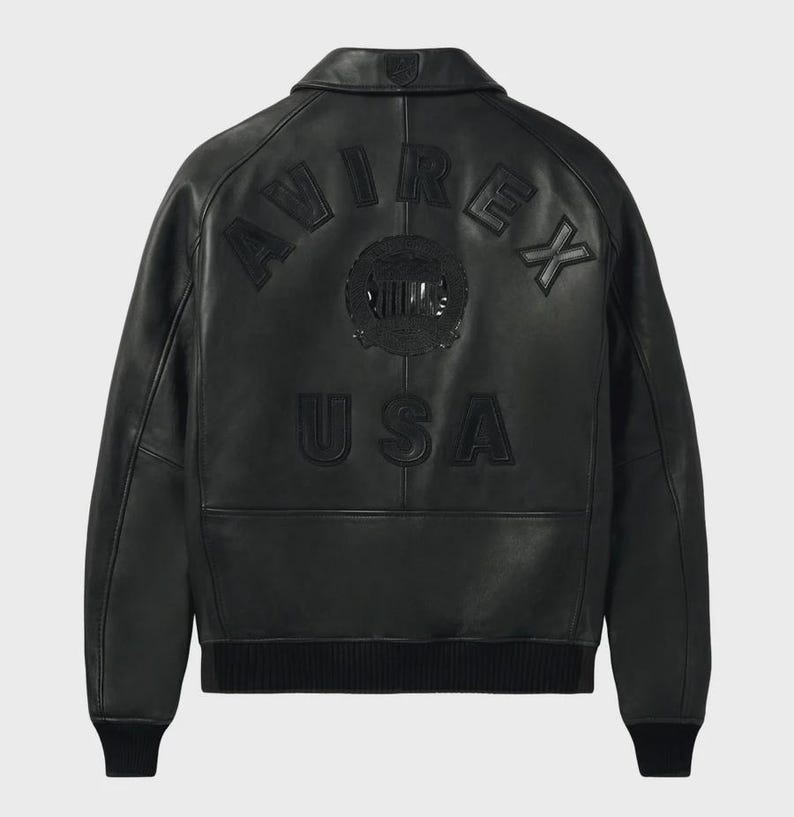 Puede incluir: Chaqueta de cuero negra con la palabra "AVIREX" arqueada en la parte superior de la espalda y "USA" debajo. La chaqueta tiene cuello, pu&ntilde;os y dobladillo de canal&eacute;. Un emblema circular est&aacute; centrado entre las palabras. La chaqueta es de estilo bomber cl&aacute;sico.